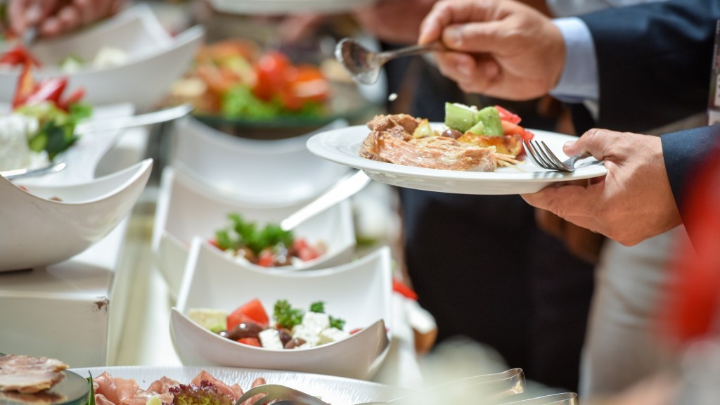 A close-up of people serving themselves from a buffet, featuring a variety of food items including salads, meats, and vegetables on white plates.