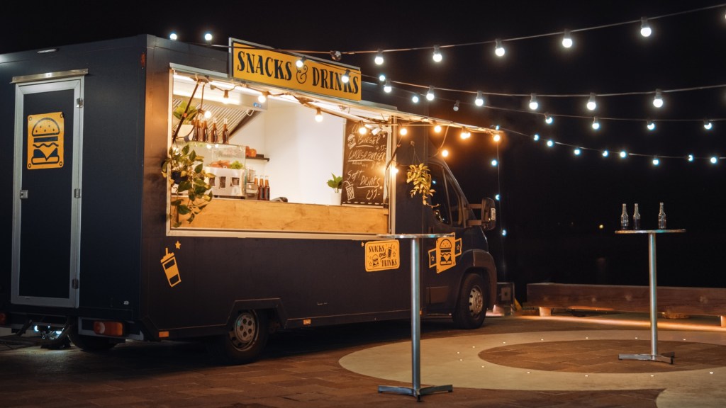 A food truck named 'Snacks & Drinks' illuminated by string lights at night, featuring a menu board and a counter with drinks.