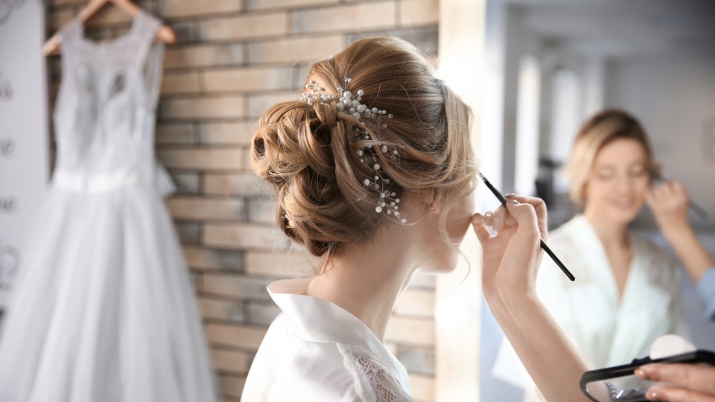 A bride getting her makeup done in front of a mirror, with her hair styled in an elegant updo adorned with hair accessories, while a wedding dress hangs in the background.