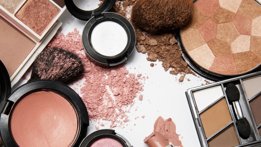An assortment of various makeup products including blush, highlighters, eyeshadow palettes, and a makeup brush, scattered on a white surface with some loose powder.